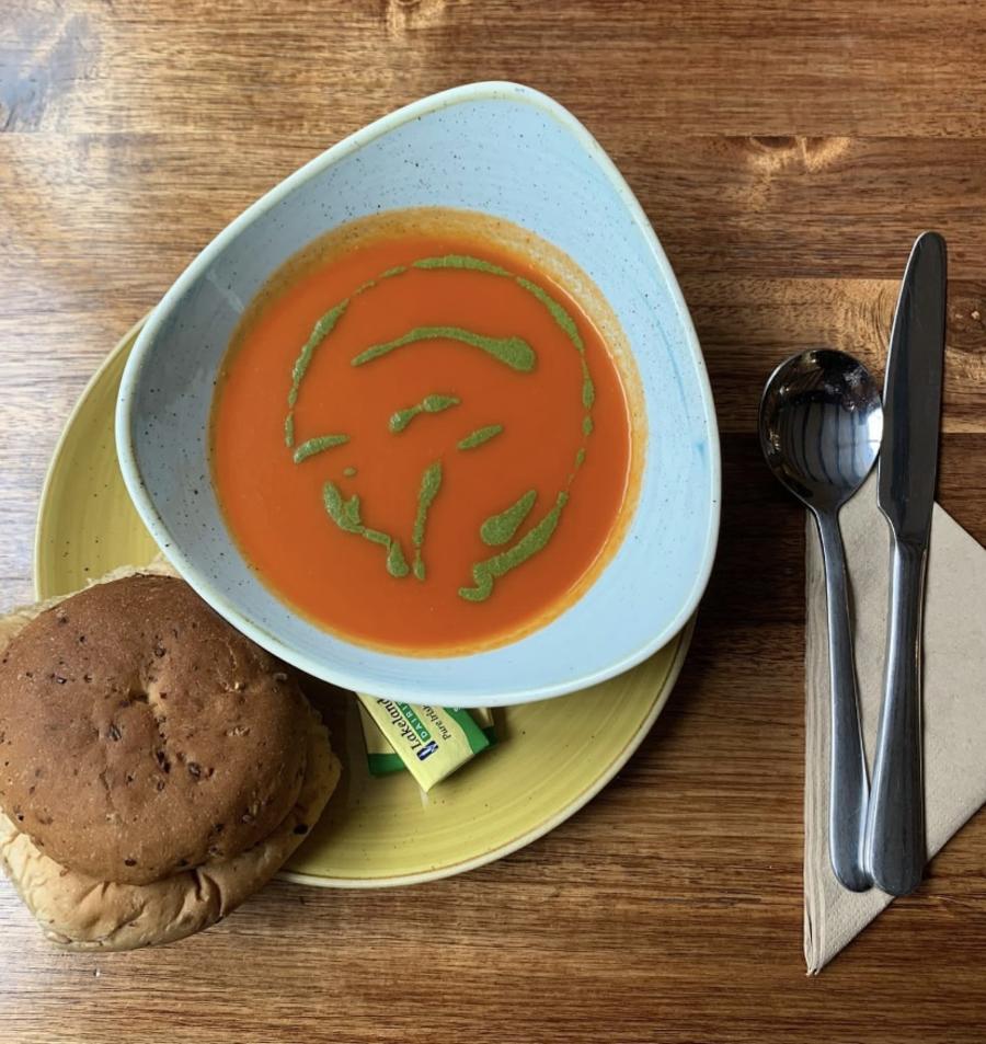 The Farrier - Wonderful Winchcombe Overhead view of a place setting on a wooden table featuring a bowl of smooth tomato soup drizzled with green herb oil. The bowl sits on a light green plate, accompanied by a bread roll. To the right are a neatly placed knife and fork on a folded napkin, along with a small condiment container. The scene is warmly lit, highlighting the textures of the wood and the vibrant color of the soup.