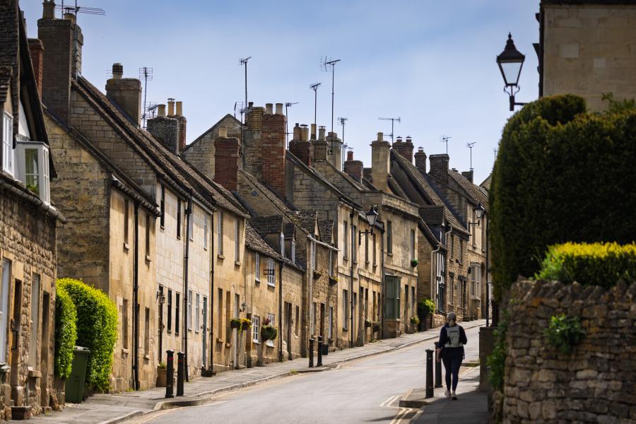 The Farrier - Wonderful Winchcombe A narrow, gently curving street lined with closely packed stone houses featuring sloped roofs, chimney stacks, and small windows. The buildings vary in warm neutral tones like beige, cream, and light brown, with some greenery climbing walls and hedges along the edges. A single pedestrian walks along the pavement on the right side of the road. A low stone wall and neatly trimmed bush appear in the foreground, while traditional street lamps rise above the homes. The sky overhead is pale and clear, casting soft daylight across the quiet scene in Winchcombe.