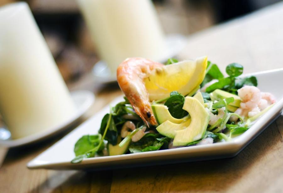 The Farrier - Wonderful Winchcombe White rectangular plate holding a composed salad with sliced avocado, leafy greens, and pieces of seafood, accompanied by a lemon wedge. The dish is placed on a wooden table, with a softly blurred background featuring a white candle and dining setting, creating a warm restaurant atmosphere.