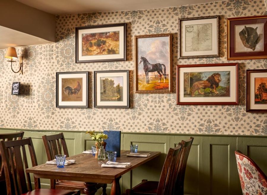 The Farrier - Wonderful Winchcombe Inside the cosy restaurant at The Lion Inn. A wooden table set for four, surrounded by dark wooden chairs. The wall behind features patterned wallpaper and is decorated with a gallery of framed artwork, many depicting horses and animals in various styles and sizes. A small vase of yellow flowers sits on the table alongside menus, plates, and cutlery. Warm, ambient lighting creates an inviting, traditional atmosphere.